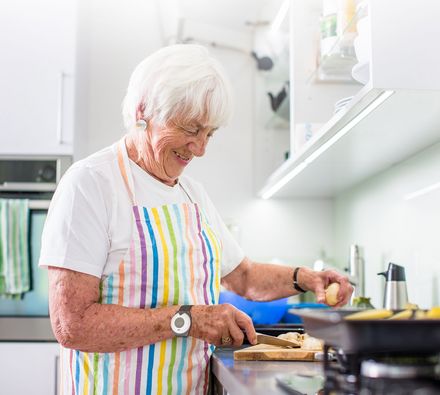Eine Seniorin steht in der Küche am Herd und trägt ihren Hausnotruf Sender am Handgelenk